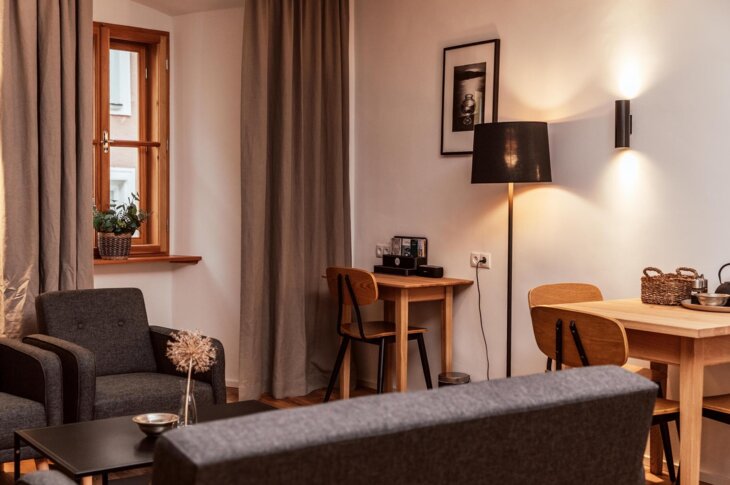 Gemütliches Wohnzimmer eines Apartments mit grauen Sitzmöbeln, Holztisch mit Stühlen und dezenter Beleuchtung.