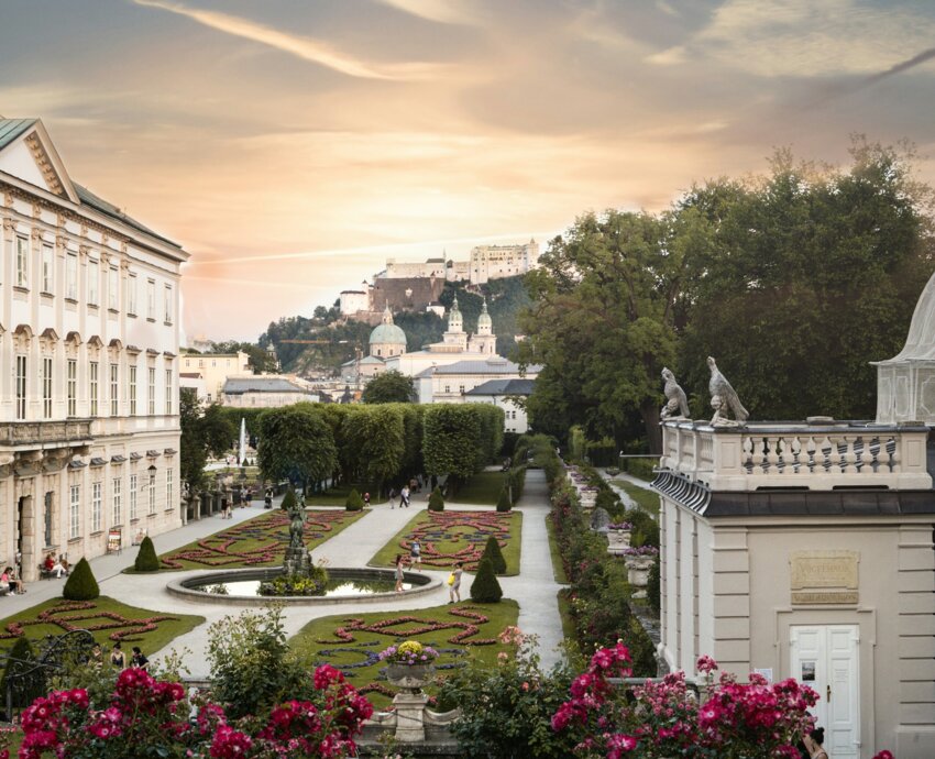 Three people sit on grass near vibrant rose bushes, overlooking Mirabell palace and garden and the historic fortress under a dramatic sky at sunset.