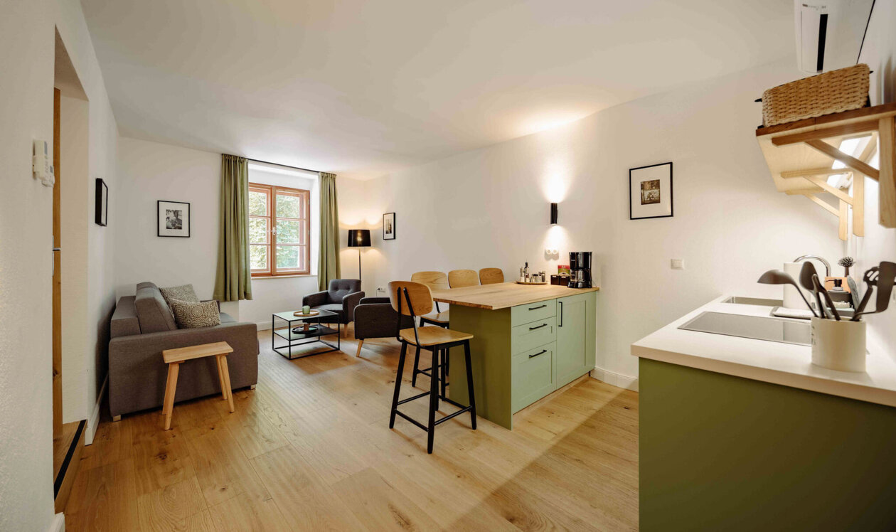 Modern living area with a cozy sitting space, wooden flooring, and a small kitchen with light green cabinets in a well-lit room.