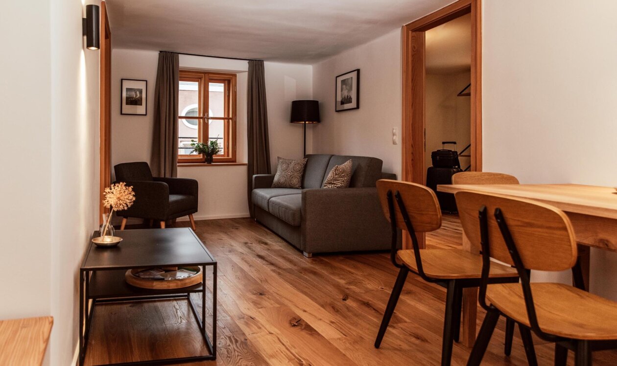 Cozy living area with wooden flooring, a grey sofa, armchair, and dining table, featuring warm lighting and a window with curtains.
