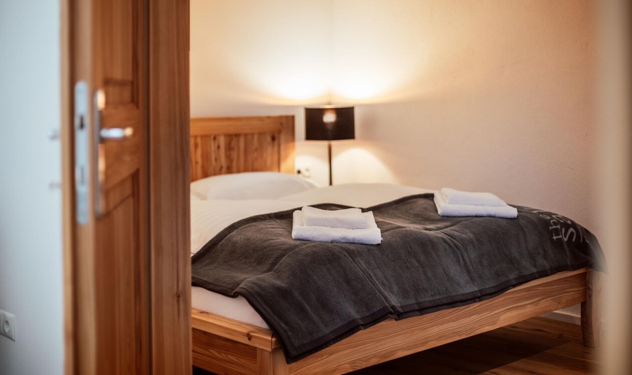 A cozy, warmly lit bedroom with a wooden bed, white bedding, and a dark blanket, viewed through an open door.