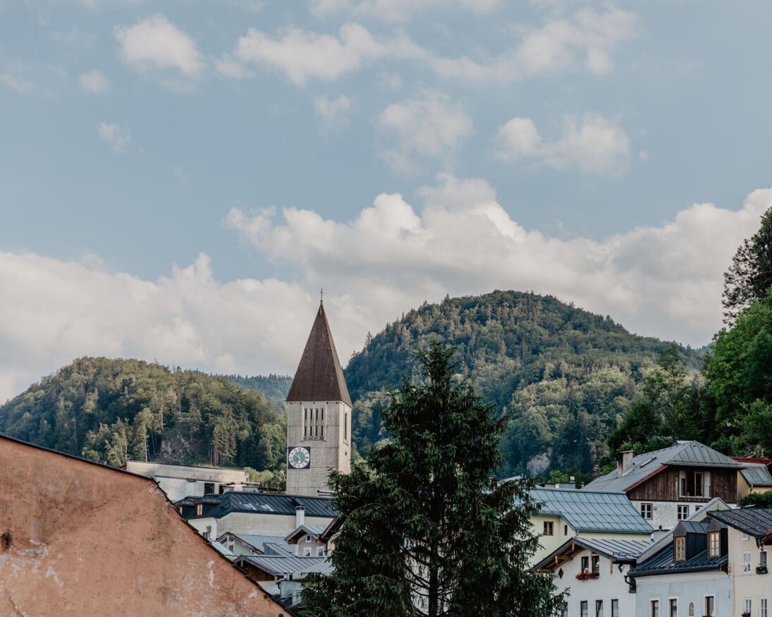 Blick auf die Stadt Hallein mit Kirchturm vor bewaldeten Hügeln und blauem Himmel.