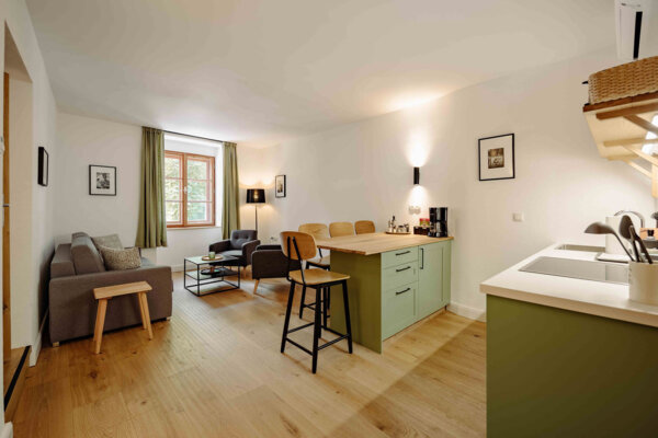 Modern living area with a cozy sitting space, wooden flooring, and a small kitchen with light green cabinets in a well-lit room.