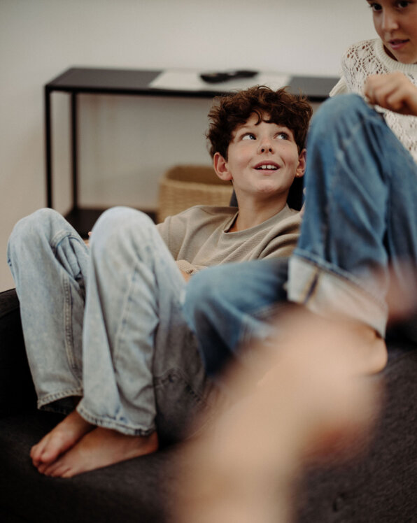 Kind sitzt entspannt auf dem Sofa und blickt zu einem Mädchen; gemütlicher Wohnbereich für Familien im Apartment von the salt in Hallein.