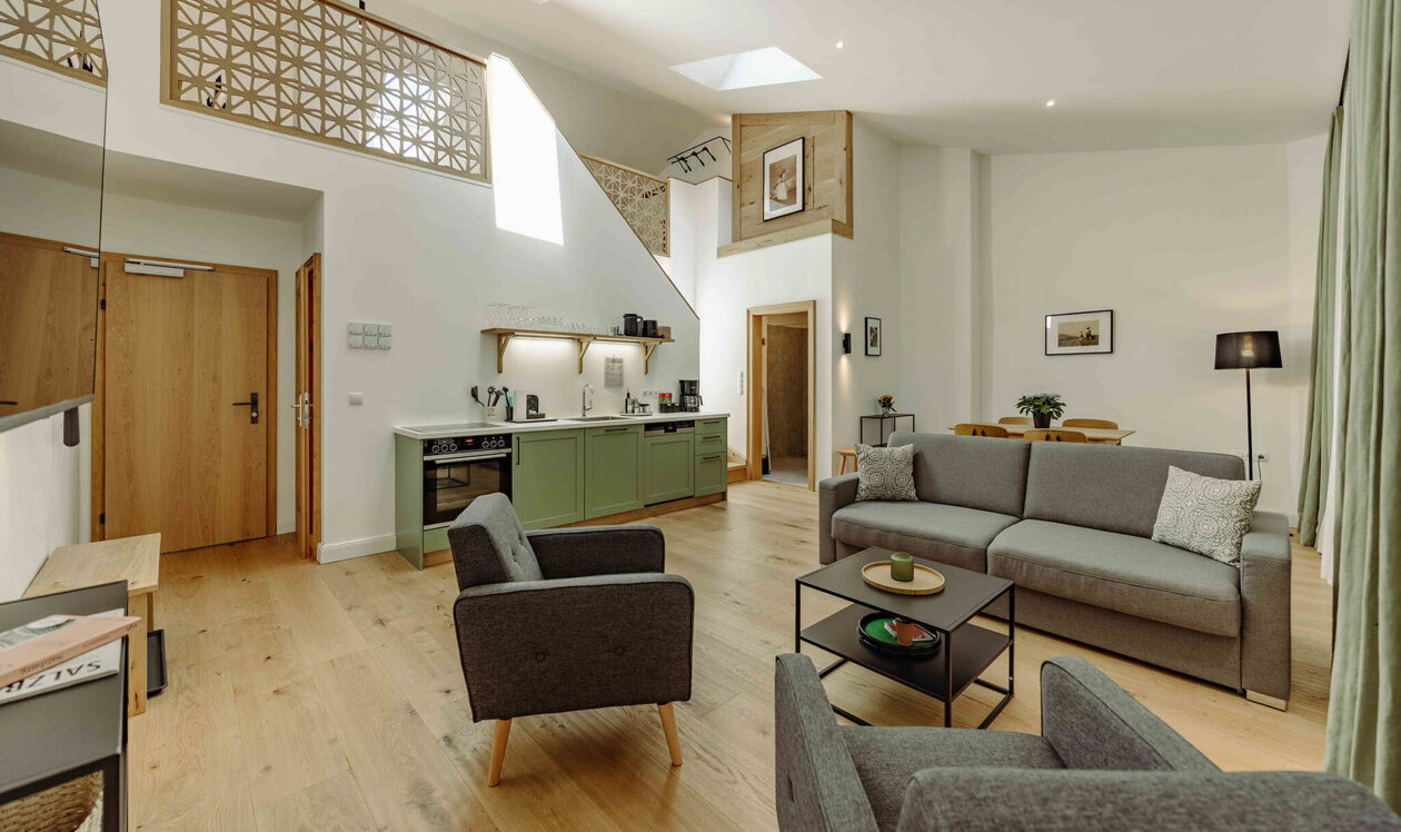 Modern living room with gray sofas, armchairs, a kitchenette with green cabinets, and decorative elements under a high ceiling with skylights.
