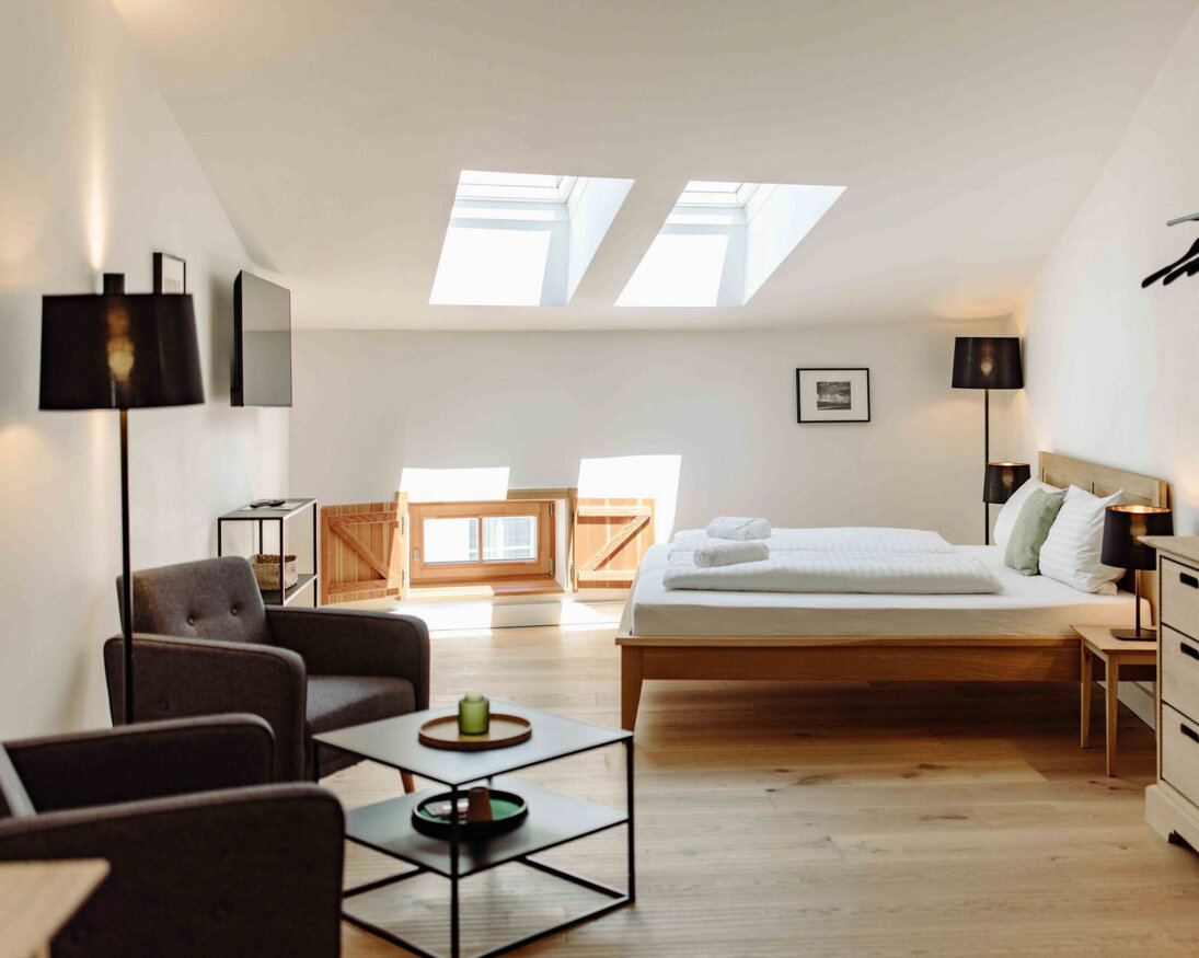 Helles, modernes Hotelzimmer mit Doppelbett, Dachfenstern, gemütlichem Sessel und Schreibtisch. Minimalistisches Design mit Holzmöbeln und Pflanzen.