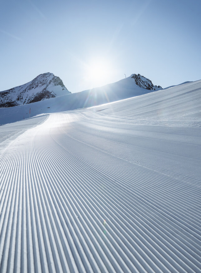 Perfekt präparierte Skipiste bei Sonnenschein, umgeben von verschneiten Bergen.