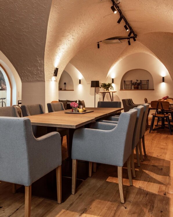 Veranstaltungsraum "Salzkammer" mit Holzboden, gewölbter Decke, großem Tisch und grauen Stühlen. Gemütliche Beleuchtung und Dekoration.