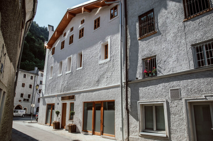 Historic white building with wooden accents on a narrow street in Hallein, featuring potted plants and a mountainous backdrop.