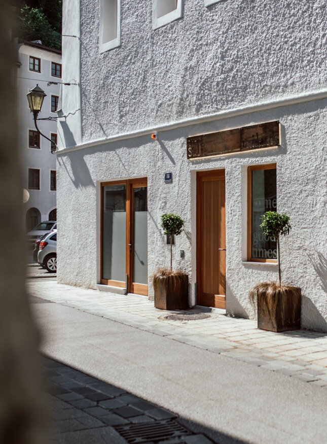 Außenansicht von the salt residences mit weißen Wänden und Holztüren, bepflanzte Töpfe vor den Eingängen, sonnige Straße im Hintergrund.