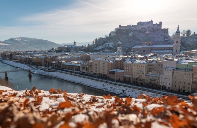 Winterlicher Blick über die Salzach auf Salzburgs Altstadt mit schneebedeckten Dächern und der Festung Hohensalzburg.