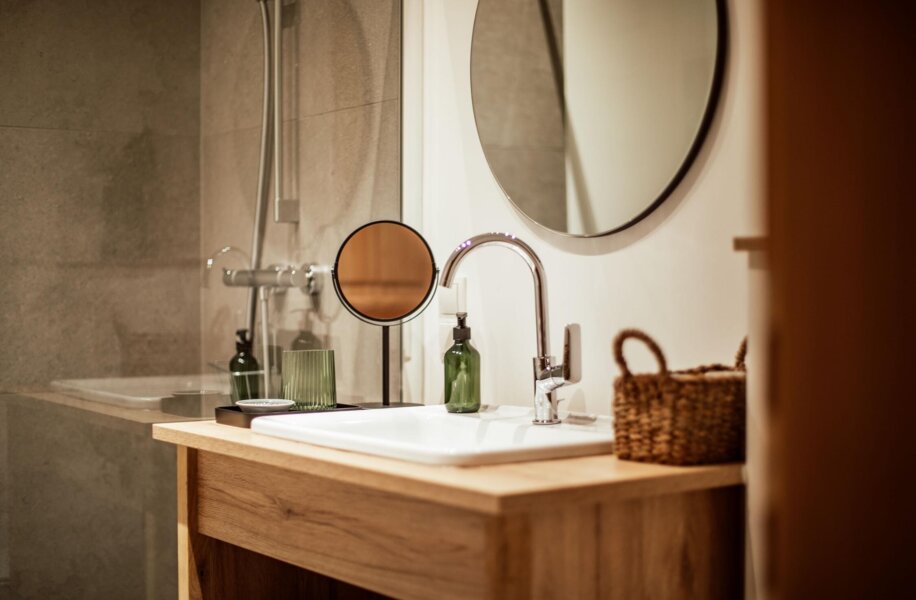 Modernes Badezimmer mit Holzwaschtisch, rundem Spiegel, Glasflaschen und Korb. Elegante Einrichtung in neutralen Tönen.