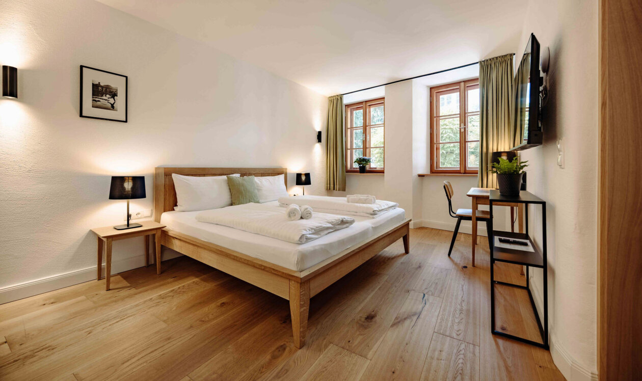 Bright hotel room with a double bed, wooden floor, and modern furnishings. Large window with curtains, desk, and flat-screen TV on the wall.