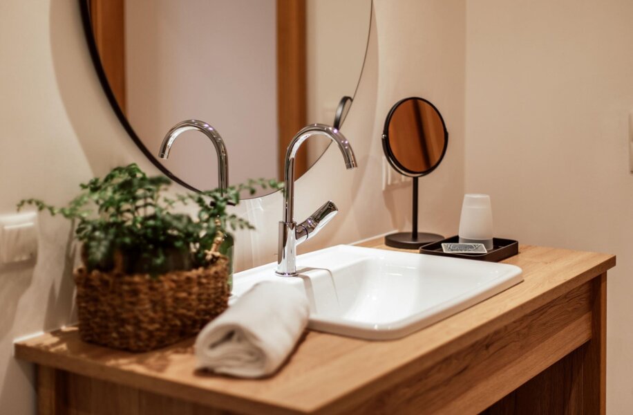 Modern bathroom sink with a round mirror, potted plant, and rolled towel on a wooden counter.