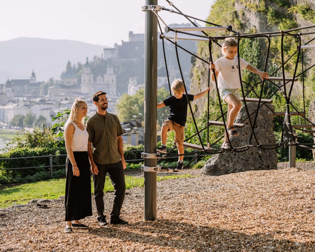 Eltern beobachten ihre Kinder beim Klettern am Spielplatz, im Hintergrund die Altstadt von Salzburg.
