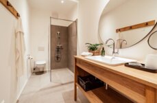 Modern bathroom with a wooden vanity, round mirror, and walk-in shower. Minimalist design featuring neutral colors and natural accents.