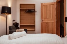 A cozy hotel room with a bed, wooden wardrobe, and open door. A floor lamp and suitcase are visible.