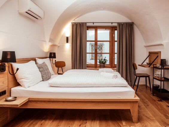 Großzügiges Hotelzimmer mit Doppelbett, Holzboden und Fenster mit Vorhängen. Moderne Einrichtung mit Schreibtisch und Getränkewagen.