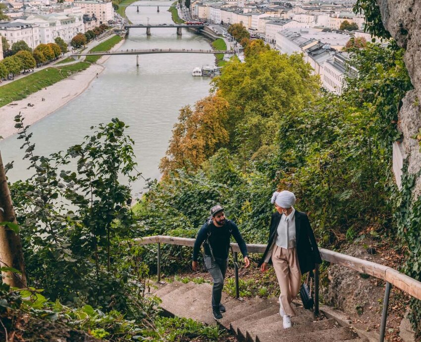 Two people walk up forested steps with a panoramic view of a riverfront city, bridges, and mountains.