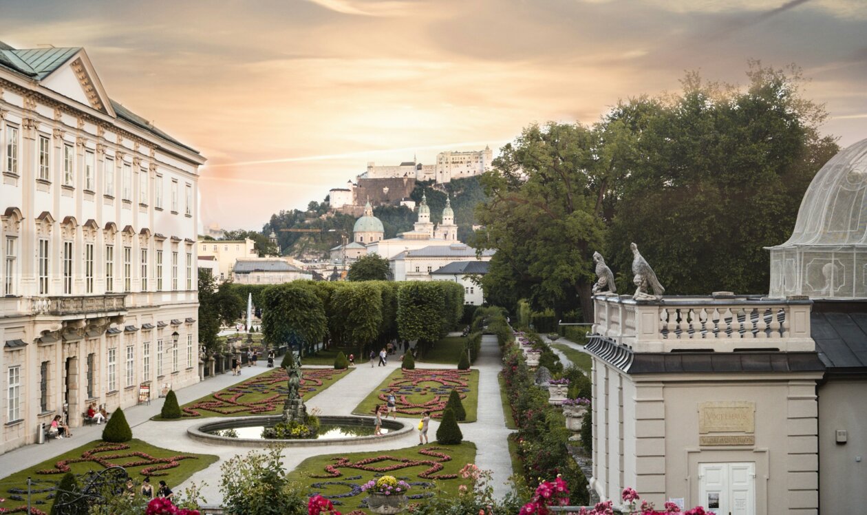 Drei Personen sitzen auf einer Wiese im Mirabellgarten, Salzburg, bei Sonnenuntergang. Im Hintergrund ist die Festung Hohensalzburg zu sehen.
