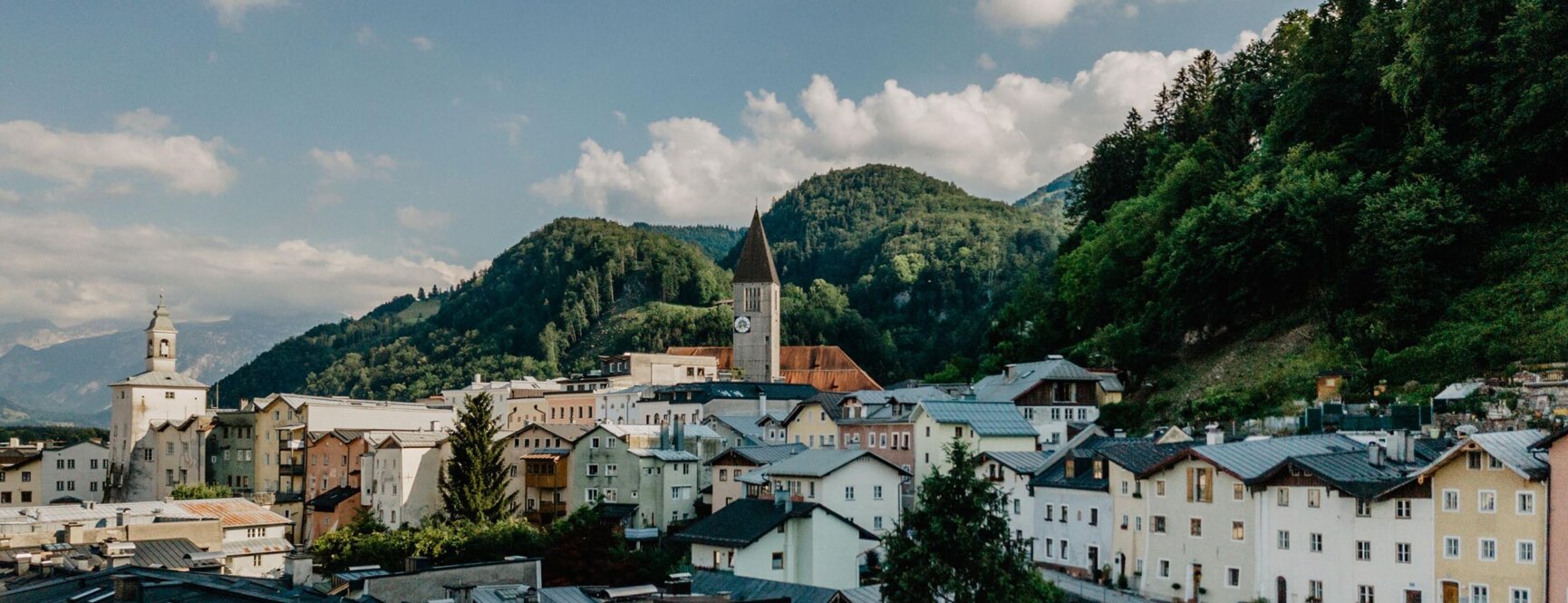 A picturesque townscape in Hallein, Austria, with colorful buildings, a church with a clock tower, and forested hills under a partly cloudy sky.