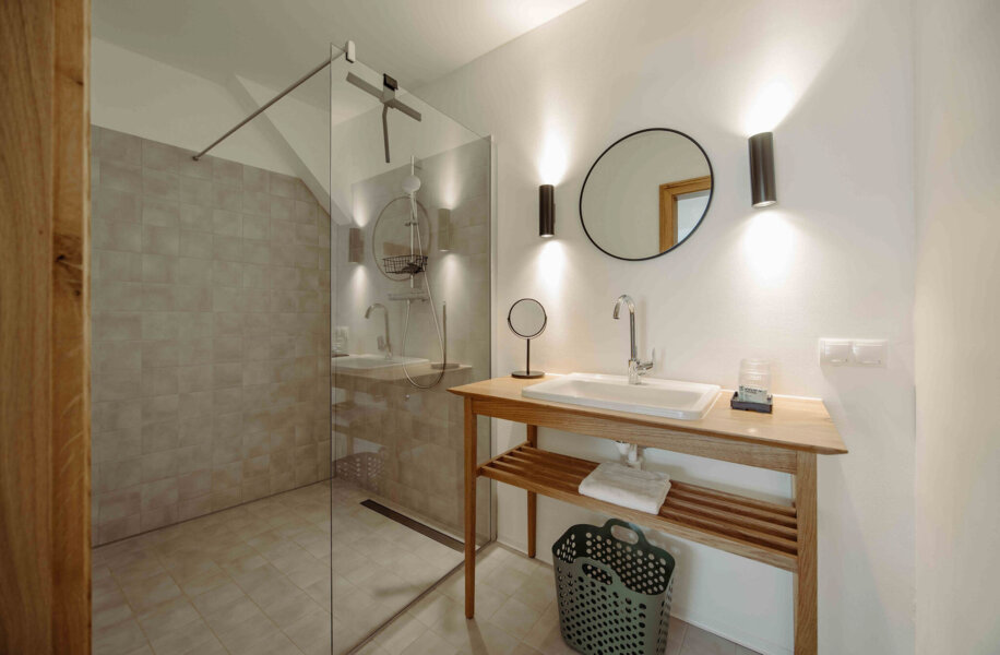 Modernes Badezimmer mit begehbarer Dusche, Holz-Waschtisch und rundem Spiegel. Dezente Beleuchtung betont die schlichte Eleganz des Raumes.