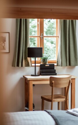 Gemütlicher Schreibtischbereich im Hotelzimmer mit Holzstuhl, Tischlampe und Fenster mit grünen Vorhängen.