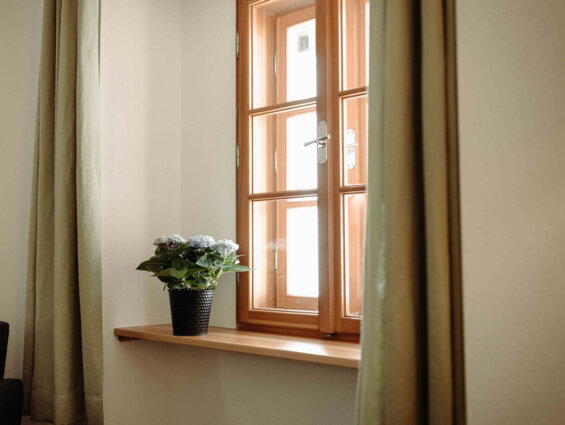 Ein Fenster mit Holzbrettern, daneben befindet sich eine Topfpflanze auf der Fensterbank und ein Vorhang.