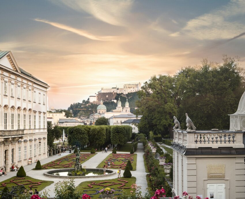 Drei Personen sitzen auf einer Wiese im Mirabellgarten, Salzburg, bei Sonnenuntergang. Im Hintergrund ist die Festung Hohensalzburg zu sehen.