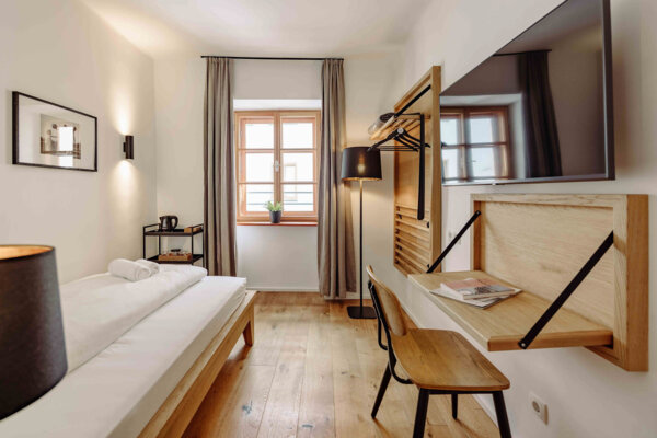 Modern hotel room with a single bed, wooden floors, a wall-mounted TV, a desk, and a window with curtains.
