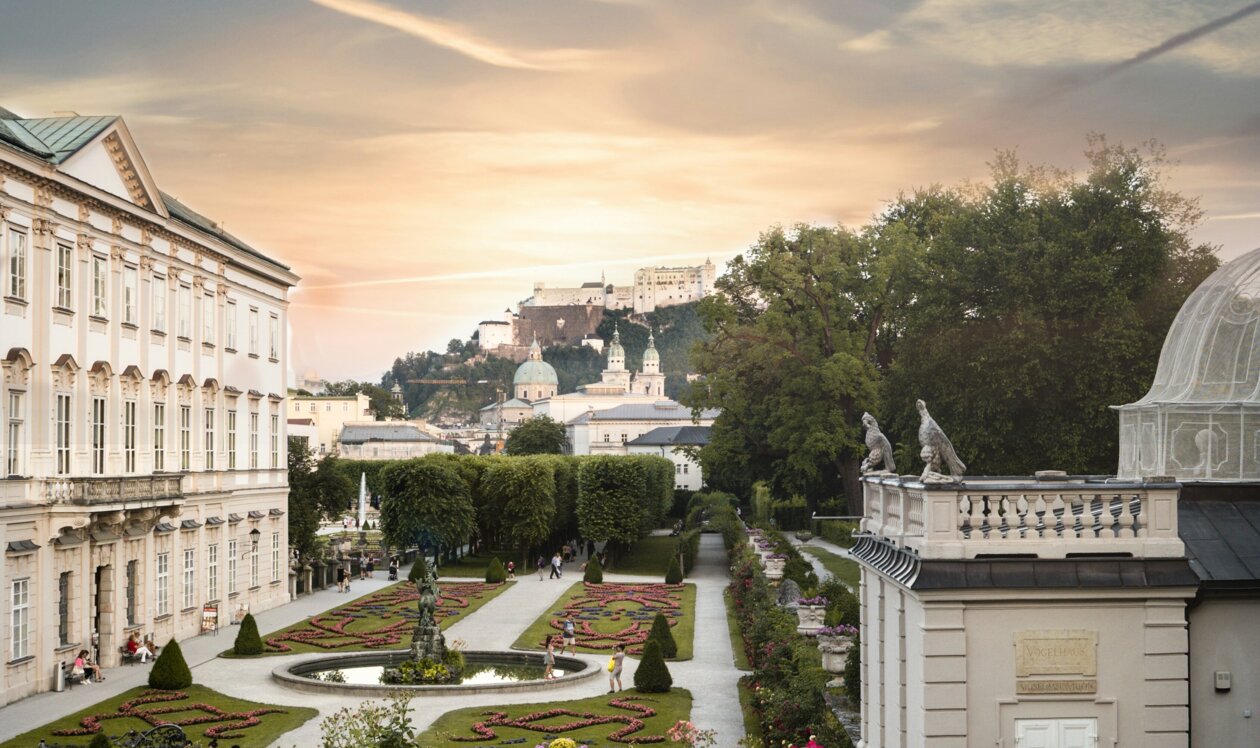 Drei Personen sitzen auf einer Wiese im Mirabellgarten, Salzburg, bei Sonnenuntergang. Im Hintergrund ist die Festung Hohensalzburg zu sehen.