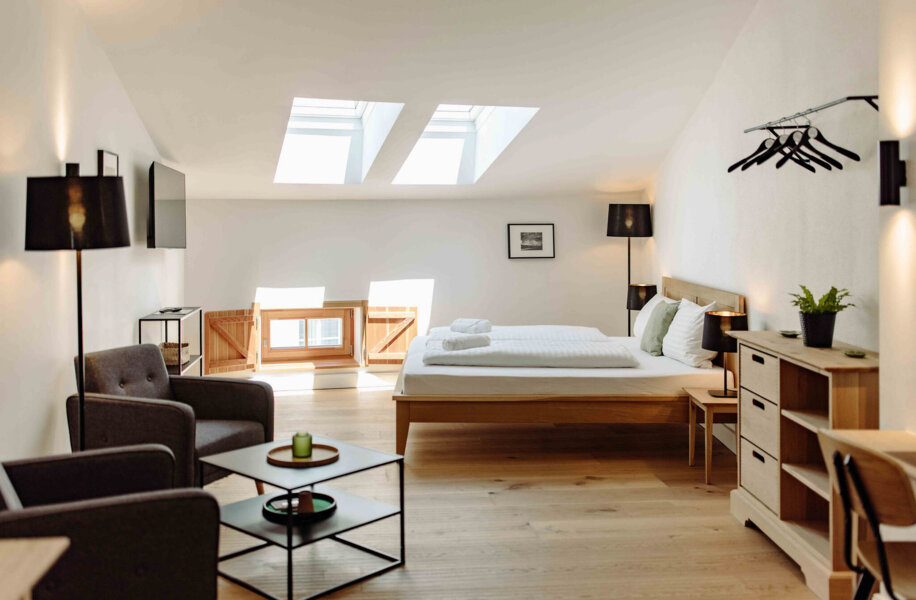 Helles, modernes Hotelzimmer mit Doppelbett, Dachfenstern, gemütlichem Sessel und Schreibtisch. Minimalistisches Design mit Holzmöbeln und Pflanzen.