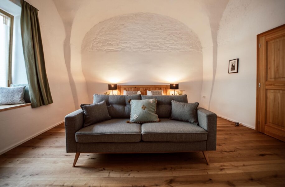 Geteilter Wohn- & Schlafbereich mit Doppelbett, grauem Sofa, Holzboden und Fensterbank. Lampen und Kissen schaffen eine einladende Atmosphäre.