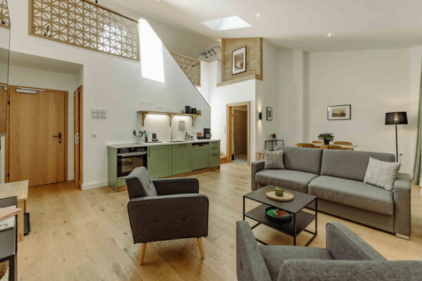 Modern living room with gray sofas, armchairs, a kitchenette with green cabinets, and decorative elements under a high ceiling with skylights.