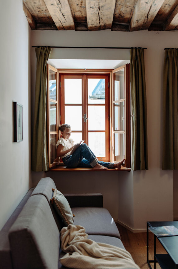 Mädchen sitzt am Fensterbrett und liest in einem hellen Zimmer mit Holzbalkendecke.