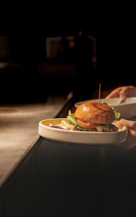 Server presenting a gourmet burger with lettuce and tomato, fries and dip on a white plate in a dimly lit restaurant.