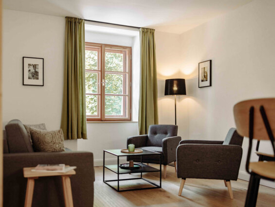 Gemütliches Wohnzimmer in einer stilvollen Ferienwohnung in Salzburg mit grauen Sesseln, Holztisch und grünen Vorhängen.
