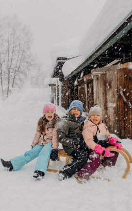 Drei Kinder auf einem Schlitten im Schnee vor einer Holzhütte, lachend und warm angezogen, erleben eine fröhliche Winterzeit.