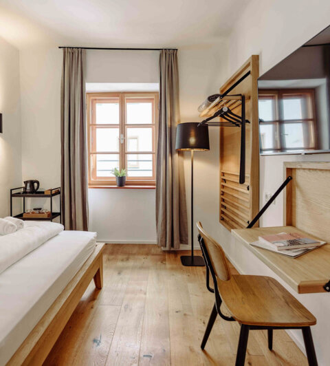 Modern hotel room with a single bed, wooden floors, a wall-mounted TV, a desk, and a window with curtains.