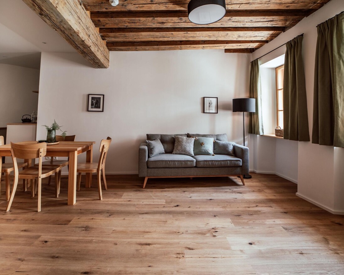 Wohnbereich mit Holzboden, grauem Sofa, Esstisch und einer voll ausgestatteten Küchenzeile. 