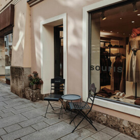 Außenansicht eines schicken Geschäfts mit zwei schwarzen Stühlen und einem kleinen Tisch auf dem Gehweg. Elegantes Schaufenster mit Kleidung.