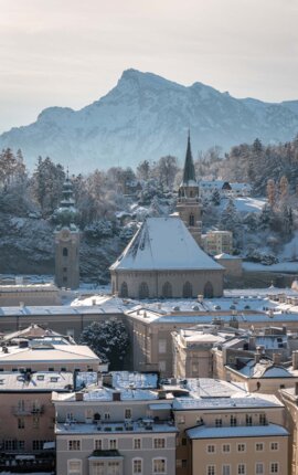 Winterlicher Blick über Salzburgs Altstadt: verschneite Dächer und Kirchtürme vor der markanten Silhouette des Untersbergs.