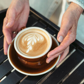 Hands holding a latte with leaf latte art in a brown cup on a black slatted cafe table.