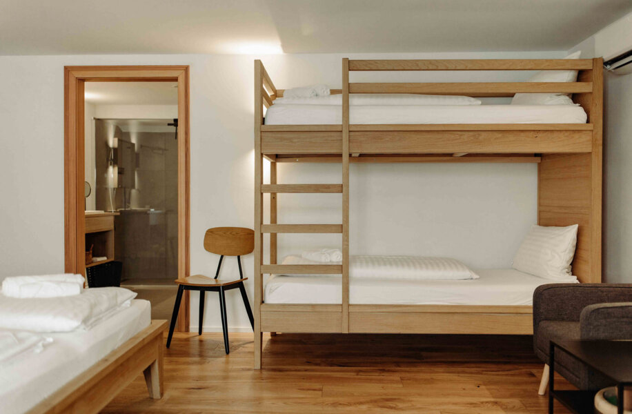 Zimmer mit Etagenbett, Holzstuhl und modernem Bad im Hintergrund.