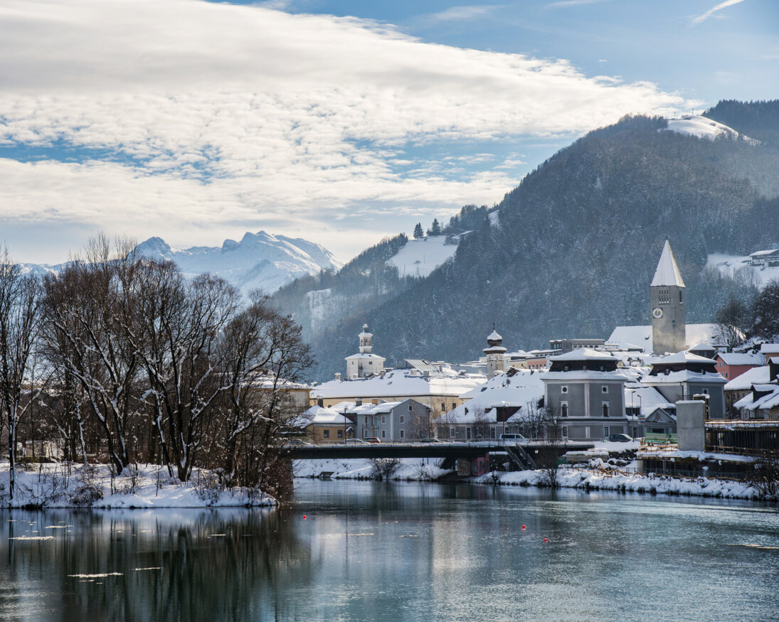 Winterliche Ansicht von Hallein: verschneite Dächer, Kirchturm und Brücke an der Salzach vor schneebedeckten Bergen.