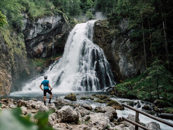 Person in Sportkleidung vor einem Wasserfall in einer grünen Waldlandschaft.