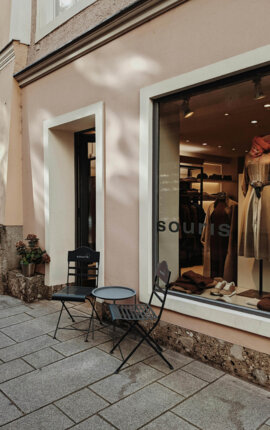Charming street view with two black chairs and a small table outside a boutique, showcasing stylish clothing in the window display.
