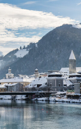 Winterliche Ansicht von Hallein: verschneite Dächer, Kirchturm und Brücke an der Salzach vor schneebedeckten Bergen.