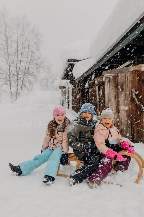 Drei Kinder auf einem Schlitten im Schnee vor einer Holzhütte, lachend und warm angezogen, erleben eine fröhliche Winterzeit.