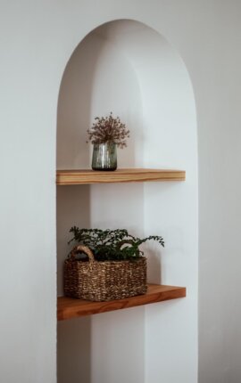 Rundbogige Wandnische mit zwei Holzregalen; oben Glasvase mit Trockenblumen, unten Flechtkorb mit Zimmerpflanze.
